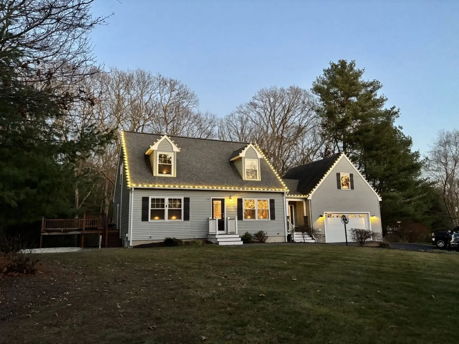 Gray cape-style home with warm white roofline lights outlining both dormers and the attached garage gable at sunset, installed by M&M Glow