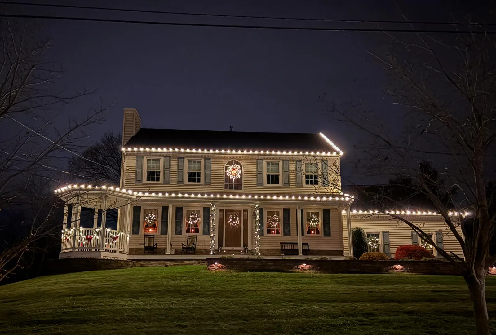 Colonial home at night with warm white C9 lights on the roofline and wraparound porch, lit wreaths in every window and wrapped porch columns, installed by M&M Glow