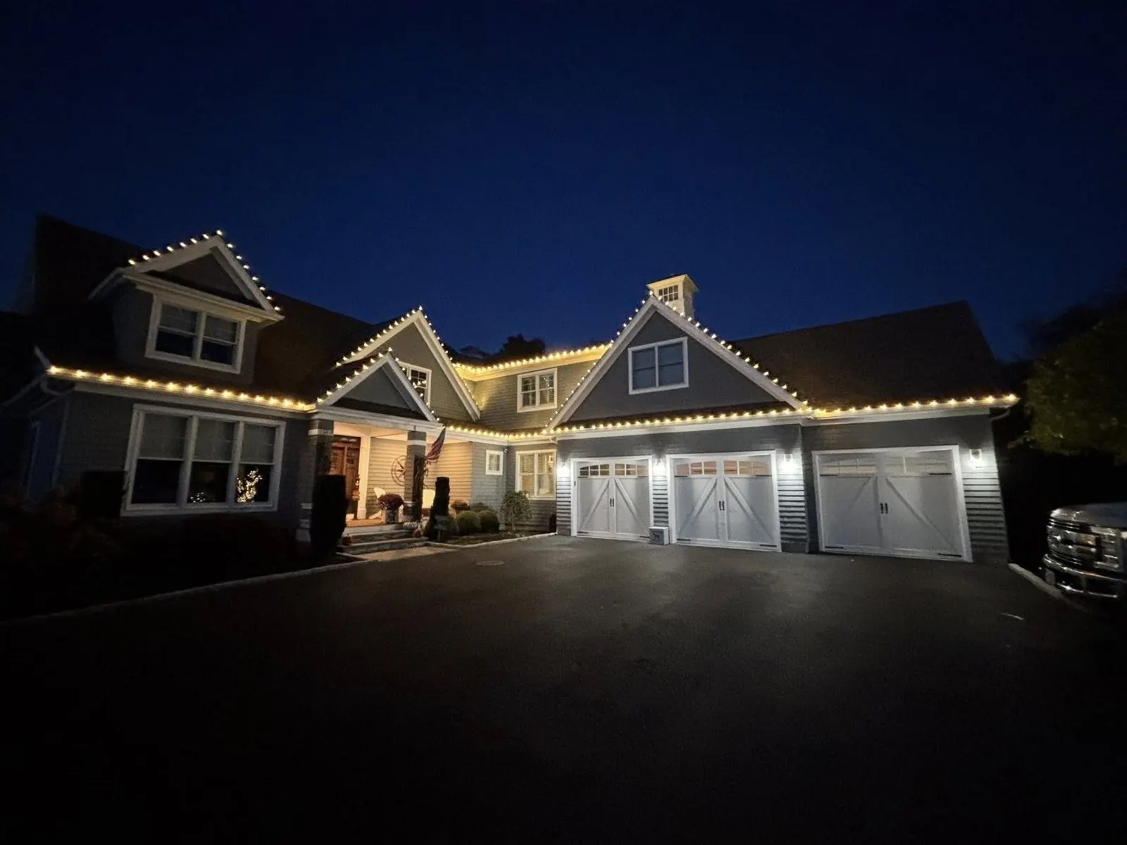 Large multi-gable home with a three-car garage outlined in warm white C9 Christmas lights along every peak and roofline at dusk, installed by M&M Glow